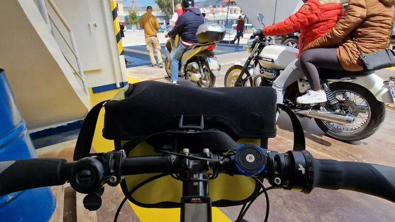 A bicycle handlebar with a yellow pannier on a ferry vehicle deck, with motorbikes and passengers behind