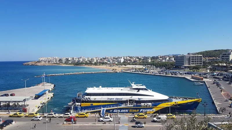 A white, blue and yellow fast ferry moored in a harbour with a beach and seafront buildings beyond