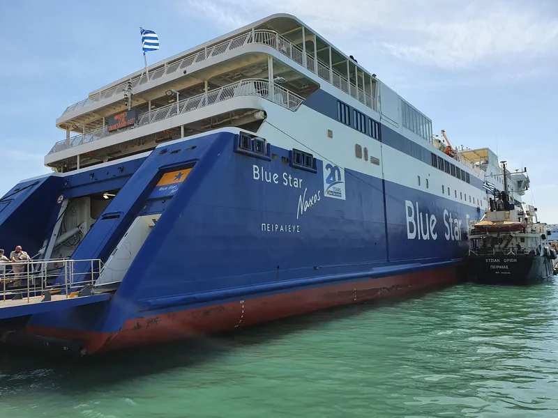 The stern of a large blue ferry moored at a quay, with its open loading ramp above green water