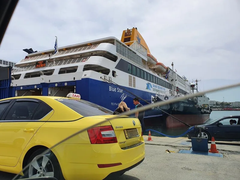 A yellow taxi waiting on the quay beside a large blue and white Blue Star ferry
